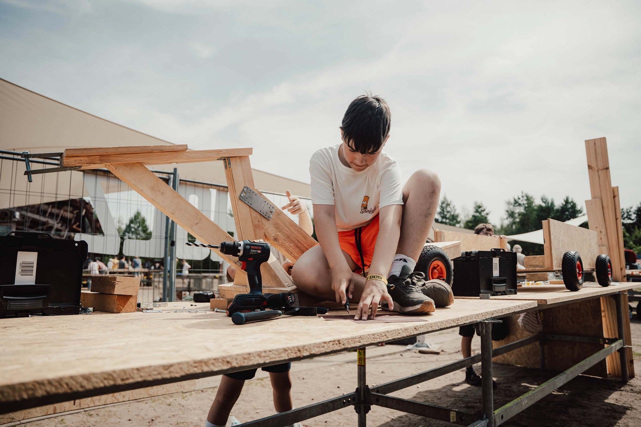 Junge arbeitet an einem Holzprojekt mit Werkzeugen wie Bohrmaschine und Schrauben.