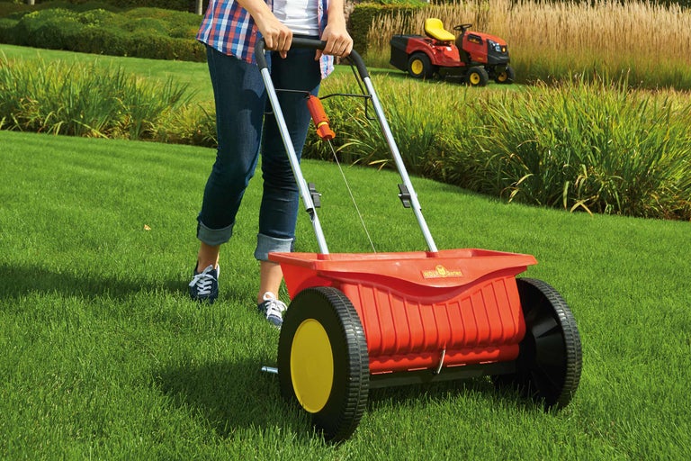 Eine Frau benutzt einen Streuwagen im Garten, im Hintergrund ist ein Rasentraktor zu sehen.