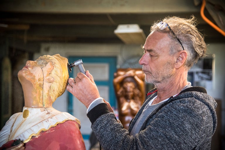 Ein Mann bearbeitet eine Holzstatue mit einem Schleifwerkzeug in einer Werkstatt.