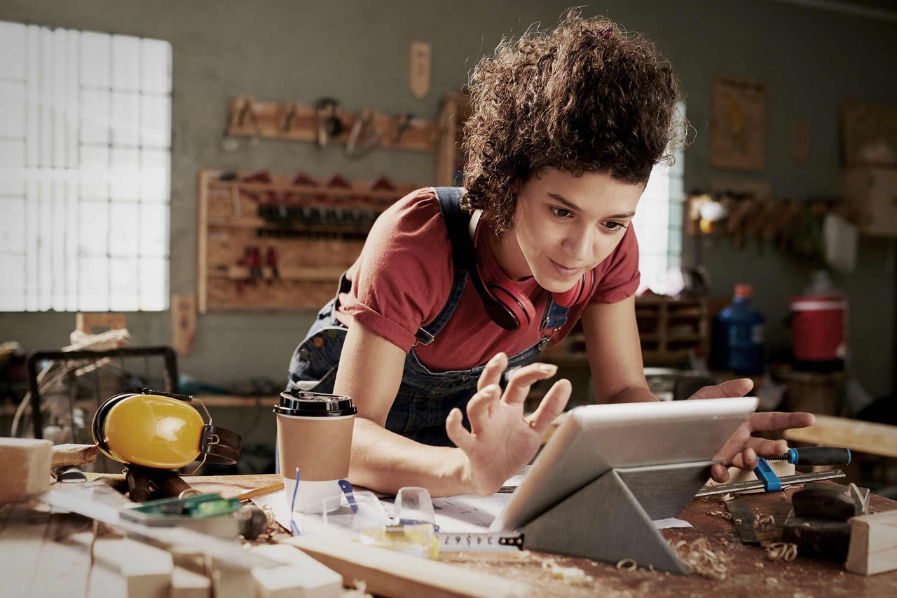 Eine Person mit lockigem Haar arbeitet in einer Werkstatt und blickt auf ein Tablet auf einer Werkbank mit Gehörschutz, Maßband und Holzstücken.