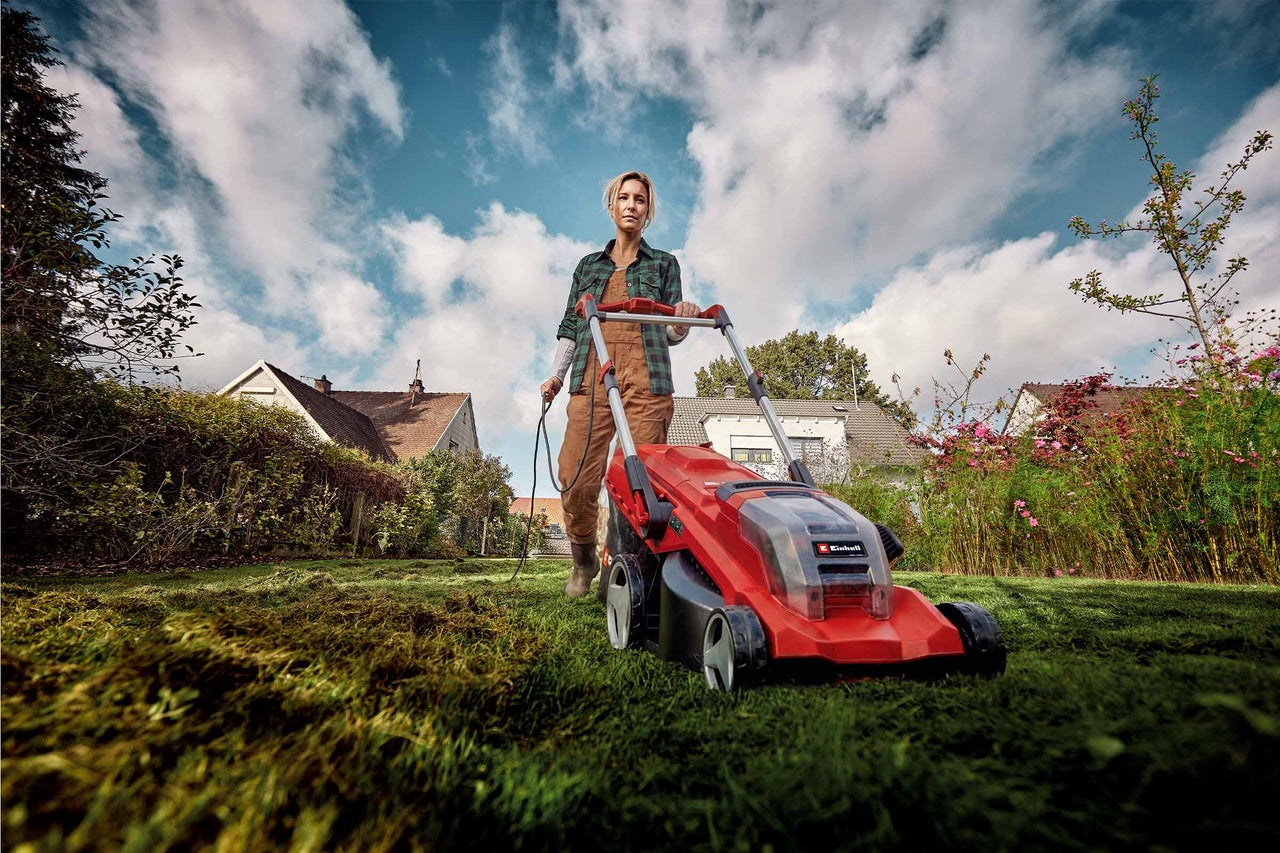 Eine Frau mäht mit einem roten Einhell Elektro-Rasenmäher einen grünen Rasen in einem Garten mit Häusern im Hintergrund.