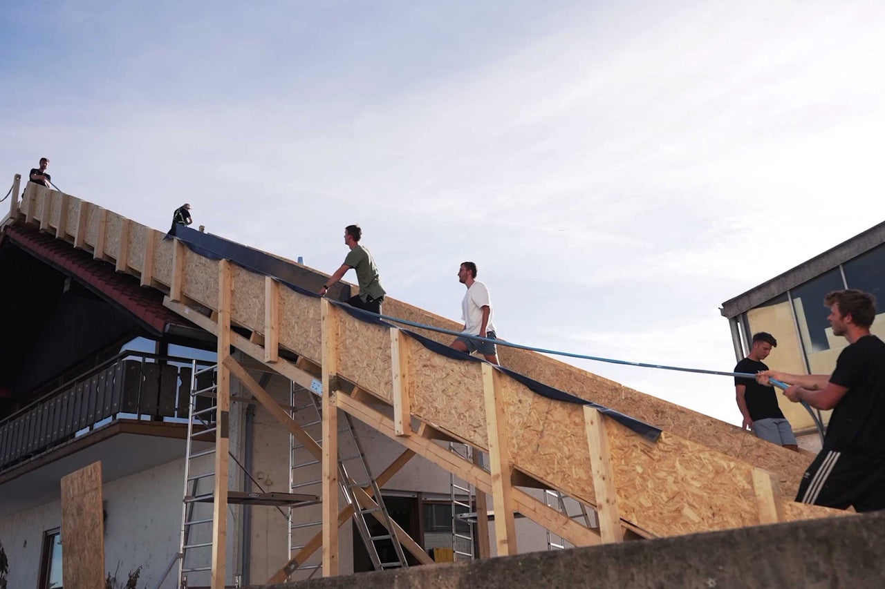 Mehrere Handwerker decken ein Dach mit Holz und Dachbahnen.