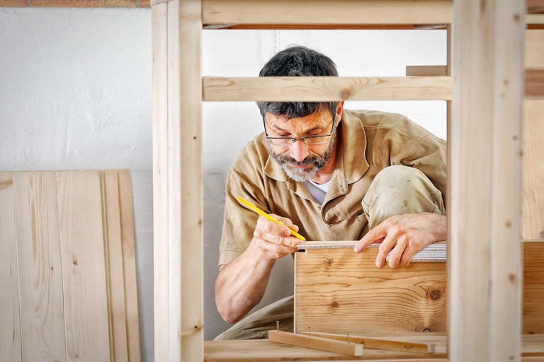 Ein Mann vermisst ein Holzbrett mit einem Zollstock und Bleistift in einer Werkstatt.