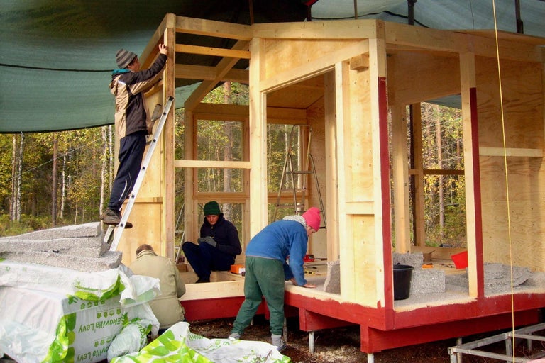 Mehrere Personen bauen ein Holzhaus mit Holzrahmen und Dämmplatten. Die Baustelle befindet sich im Freien.