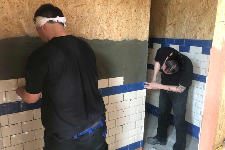 Zwei Handwerker verlegen Fliesen in einem Raum. Die Fliesen sind rechteckig und in einem Farbwechsel verlegt.