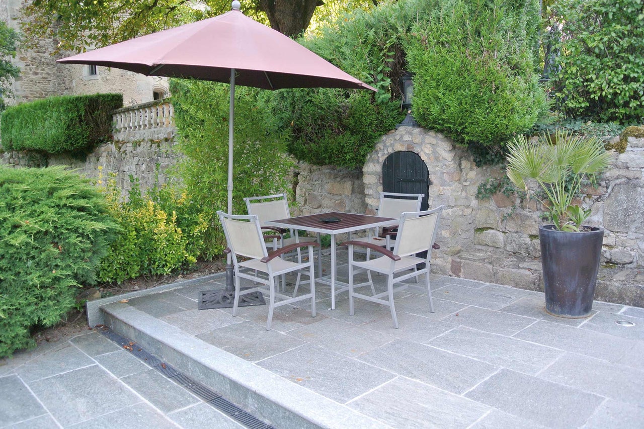 Quadratischer Gartentisch mit vier Stühlen und Sonnenschirm auf gepflasterter Terrasse