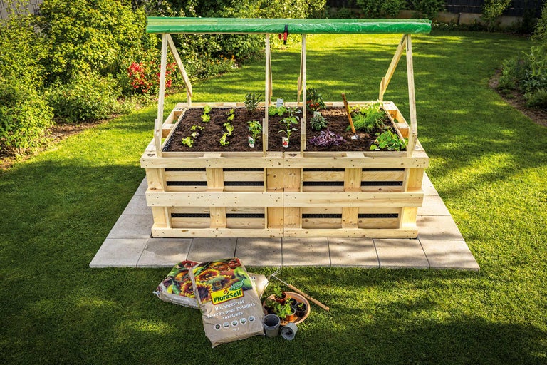 Hochbeet aus Holz mit Pflanzen und Gemüse im Garten, im Vordergrund Blumenerde.