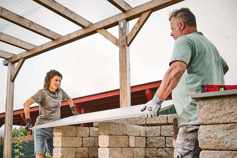 Zwei Personen errichten eine Gartenmauer mit Betonsteinen unter einem hölzernen Unterstand.