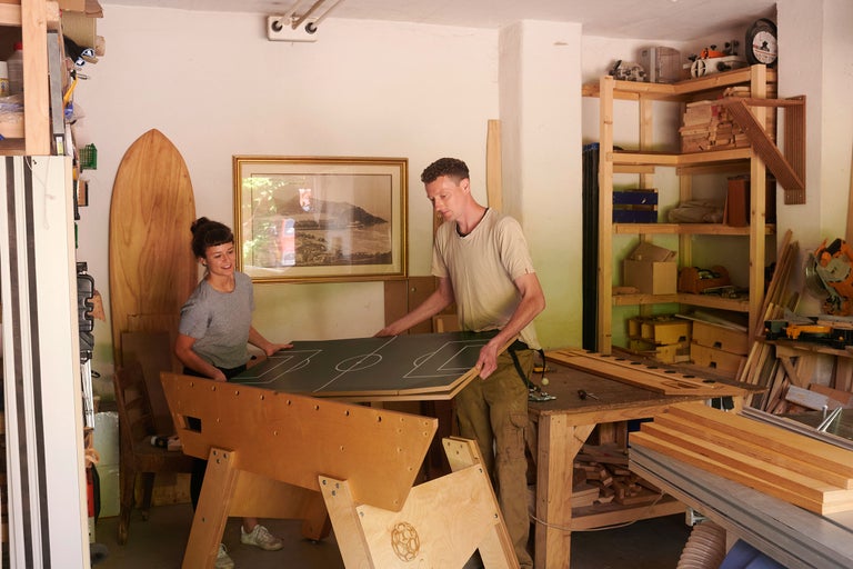 Zwei Personen montieren einen Tischfußballtisch in einer Werkstatt mit Holzregalen und Werkzeugen.