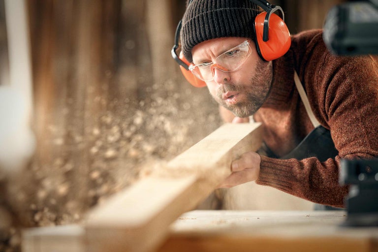 Mann bläst Sägespäne von Holzbrett, trägt Schutzbrille und Gehörschutz
