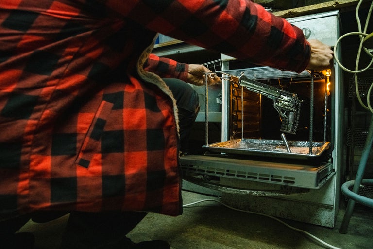 Person im rot-schwarzen Hemd, die ein Metallobjekt in einen Ofen stellt.
