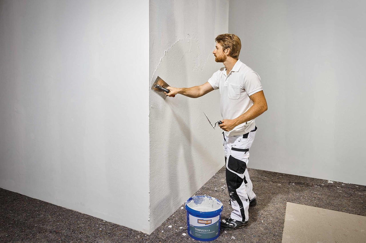 Ein Mann trägt Putz mit einer Glättkelle auf eine Wand auf, ein Eimer Putz steht daneben.