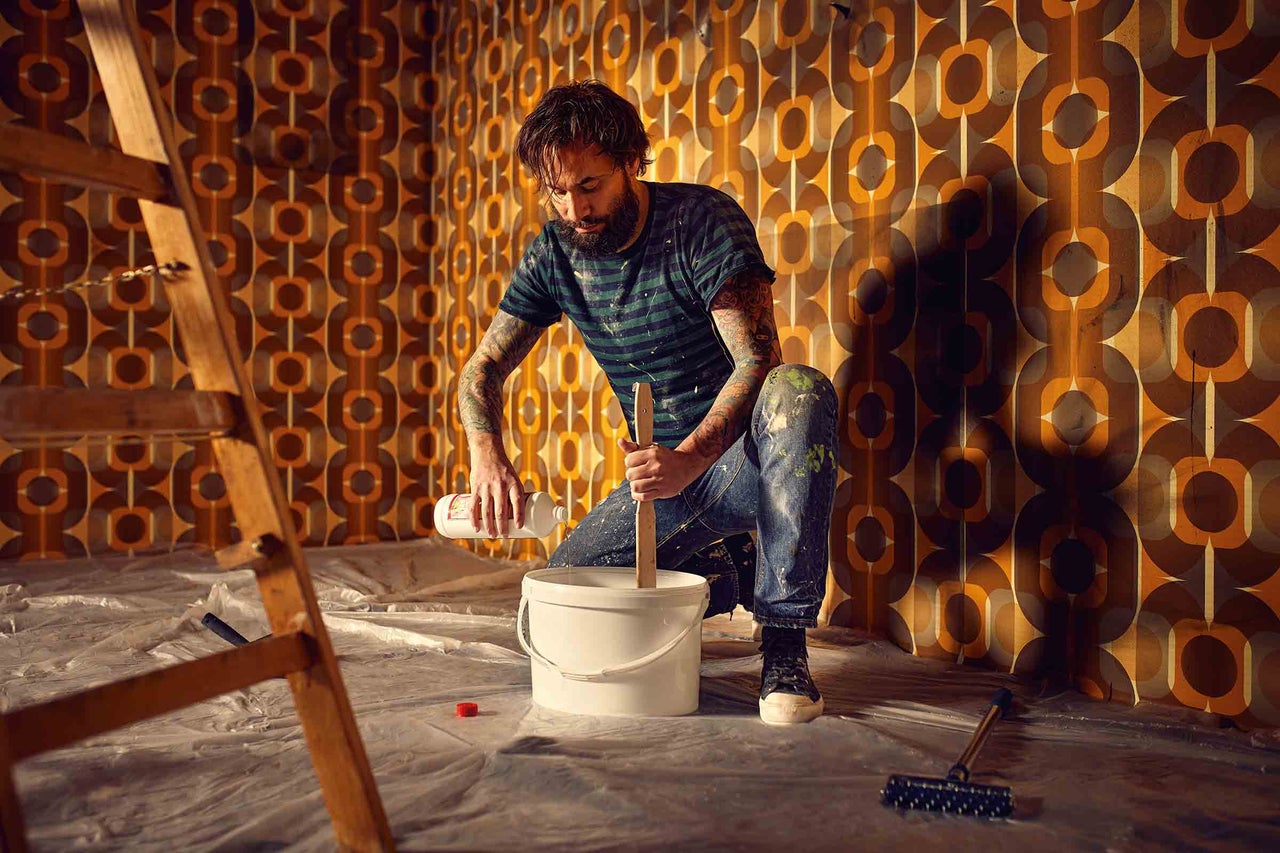 Mann mischt Farbe in einem Eimer für Renovierungsarbeiten in einem Raum mit Retro Tapete.