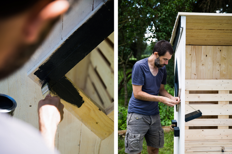Ein Mann streicht ein Holzgebäude mit schwarzer Farbe und befestigt einen Riegel.