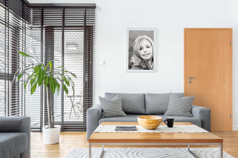 Wohnzimmer mit Couch, Tisch, Zimmerpflanze, Jalousien und einem gerahmten Schwarzweiß-Porträt an der Wand