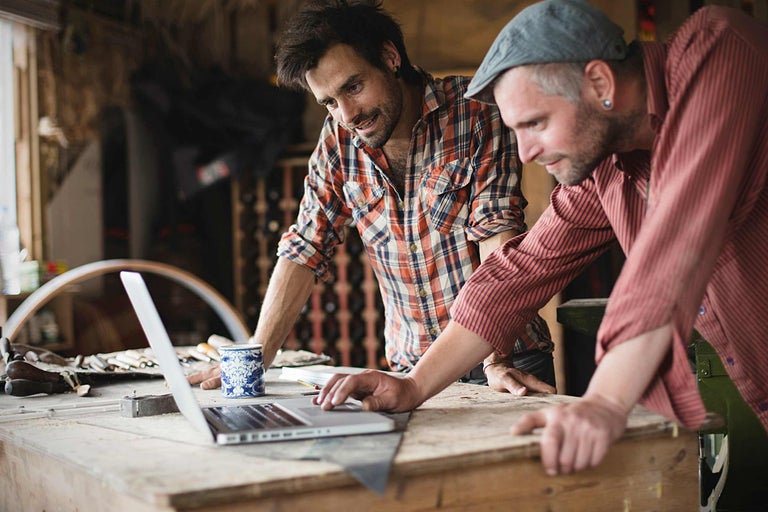 Zwei Handwerker arbeiten an einem Laptop in einer Werkstatt. Werkzeuge und Baumaterialien sind im Hintergrund zu sehen.