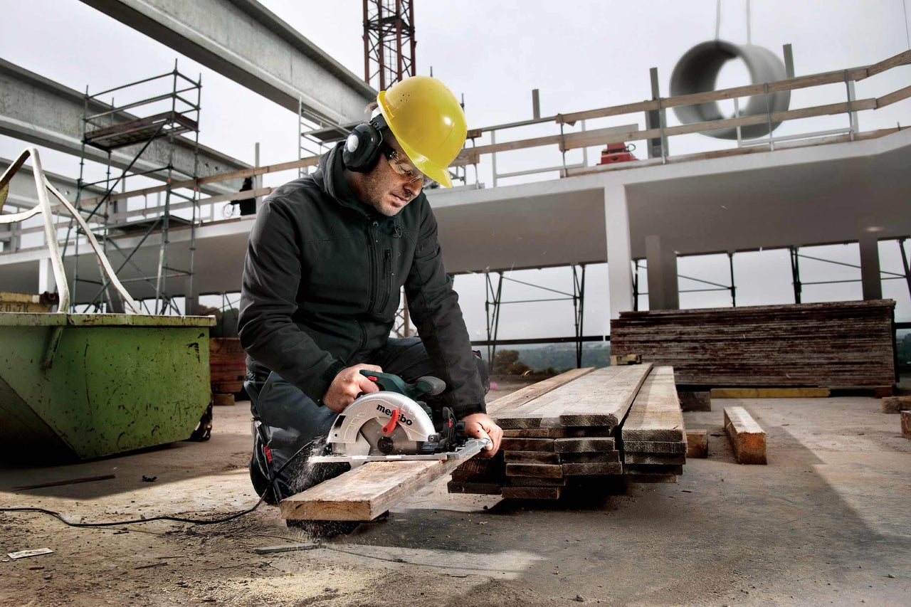 Ein Bauarbeiter mit Schutzhelm, Gehörschutz und Schutzbrille sägt ein Holzbrett mit einer Handkreissäge auf einer Baustelle.