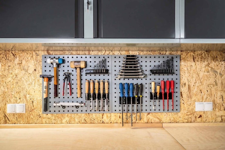 Organisierte Werkzeugwand mit grauen Lochplatten an einer OSB-Wand über einer Werkbank, mit Hämmern, Zangen, Beiteln und Schraubendrehern.