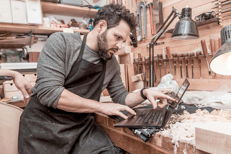 Ein Handwerker mit einer Schürze bedient einen Laptop in einer Werkstatt.