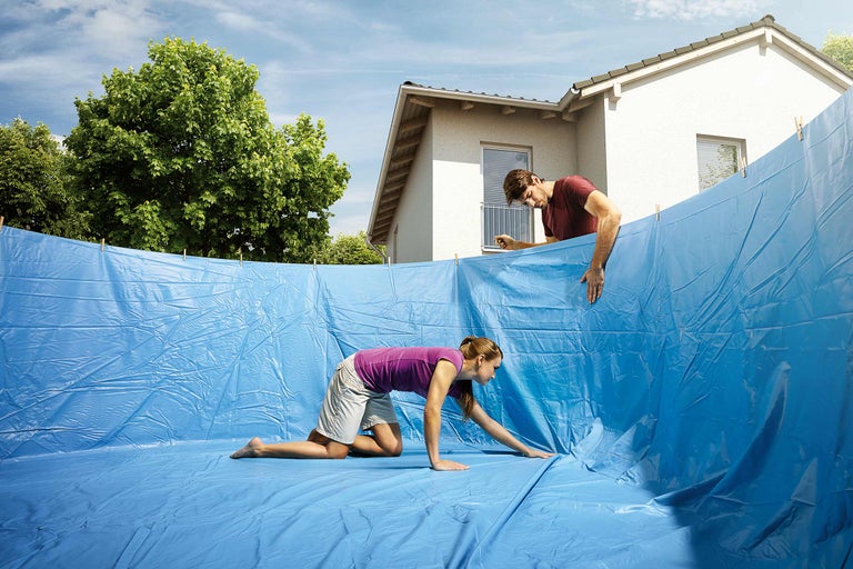 Szene mit zwei Personen, die eine blaue Poolfolie installieren, mit einem Wohnhaus und Bäumen im Hintergrund.