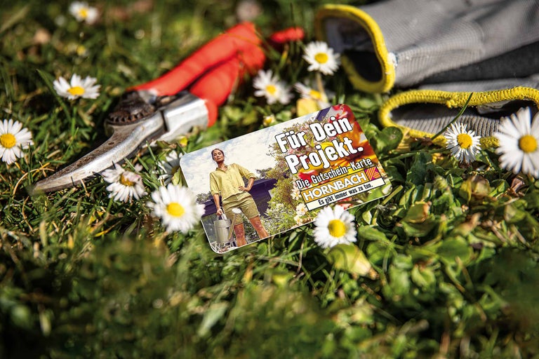 Gartenszene mit Hornbach Gutscheinkarte, Gartenschere und Gartenhandschuhen auf Gras mit Gänseblümchen