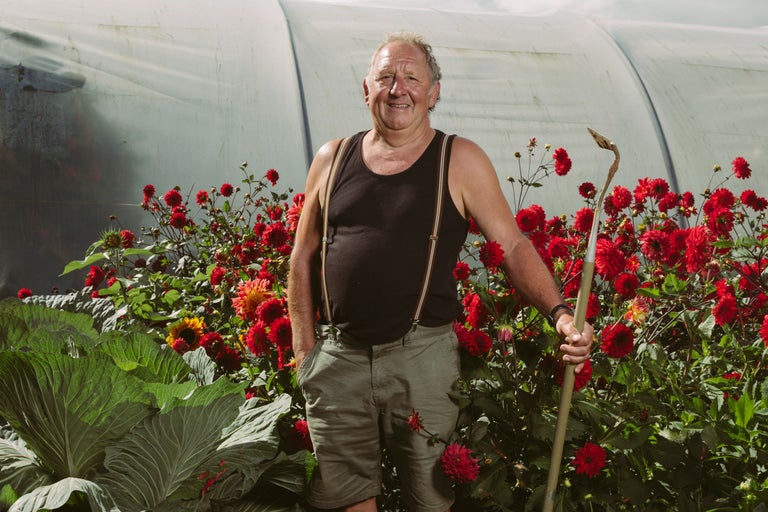 Ein Mann steht mit einer Hacke vor einem Gewächshaus, umgeben von Blumen und Gemüse.