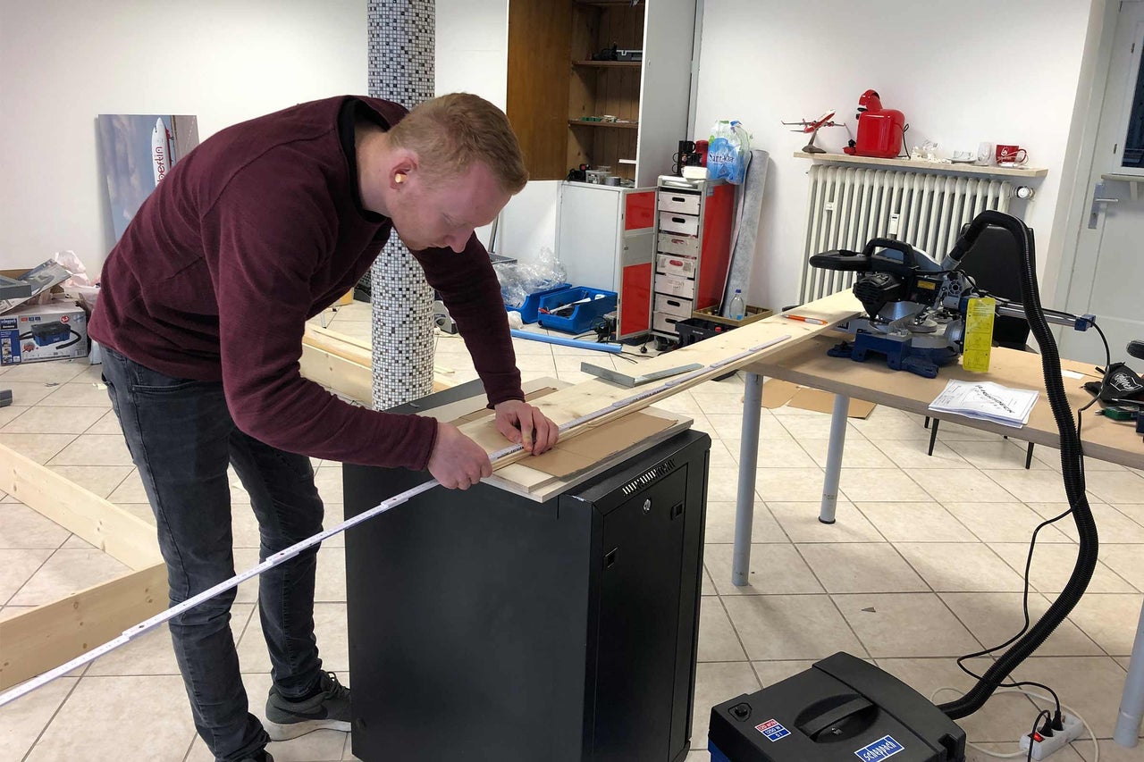 Ein Mann misst ein Holzbrett mit einem Zollstock in einer Werkstatt.