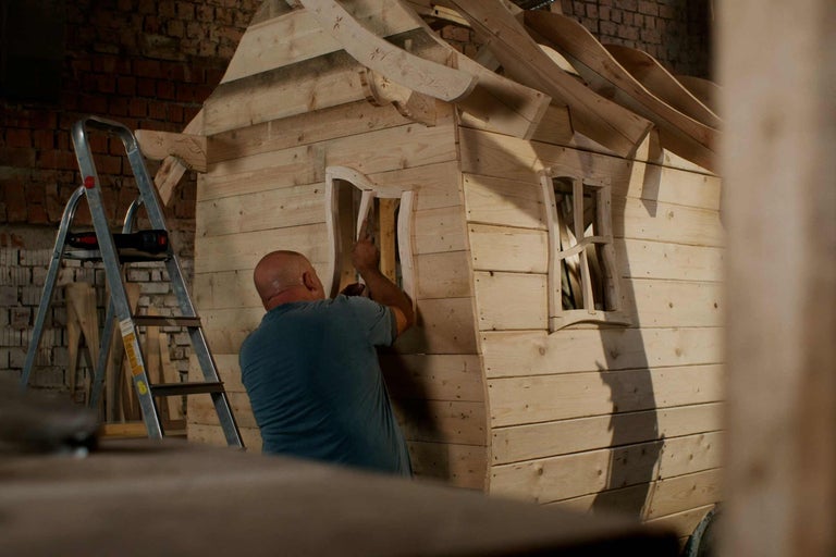 Ein Holzspielhaus wird von einem Mann gebaut. Eine Leiter steht daneben.