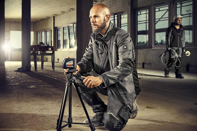 Ein Arbeiter hockt mit einem Kreuzlinienlaser und einem Stativ auf einer Baustelle.