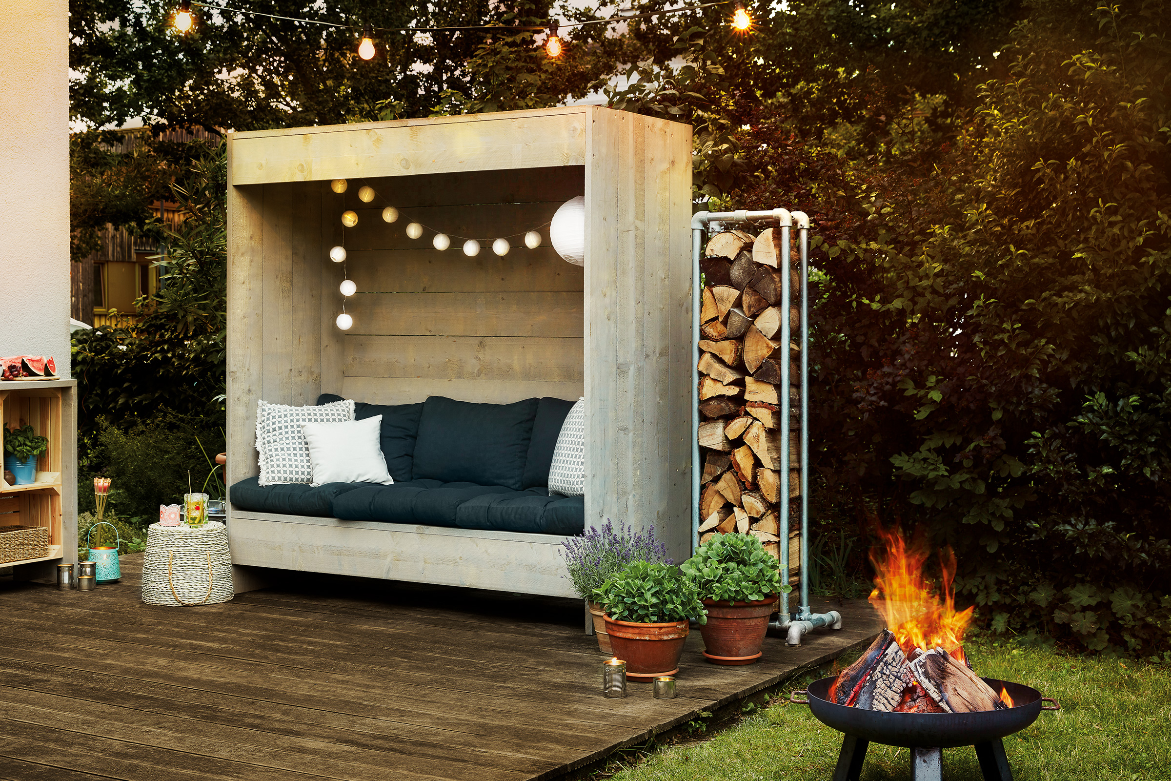 Gemütliche Gartenszene mit Holzterrasse, Sitzecke mit Kissen, Holzstapel, Blumentöpfen und Lagerfeuerstelle