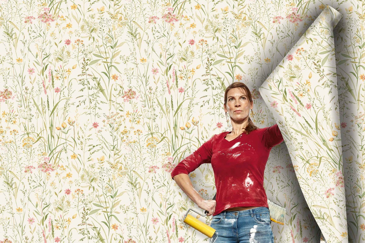 Eine Frau mit Tapezierwerkzeug steht vor einer Wand, die mit einer Vliestapete mit floralem Blumenwiesenmuster tapeziert wird.