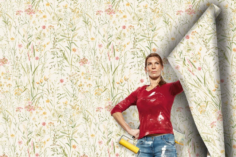 Eine Frau mit Tapezierwerkzeug steht vor einer Wand, die mit einer Vliestapete mit floralem Blumenwiesenmuster tapeziert wird.