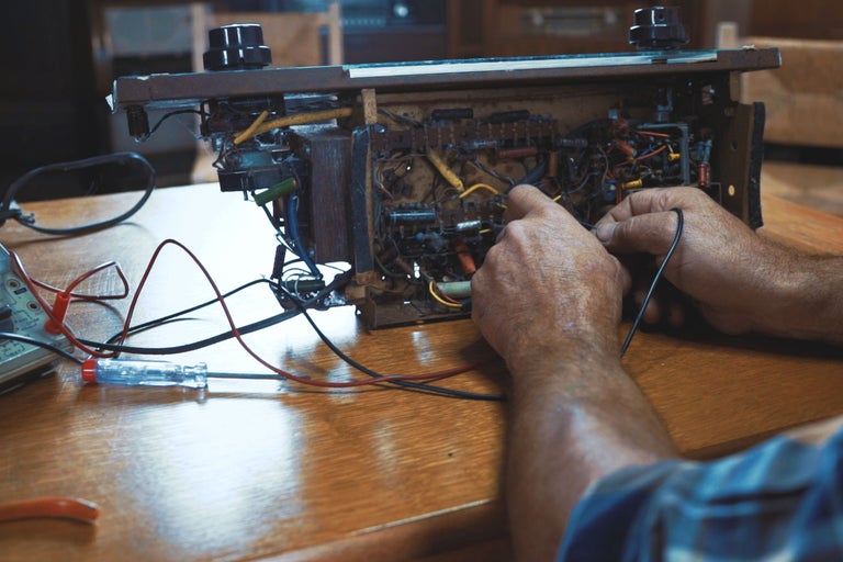 Ein Mann repariert ein Radio mit einem Schraubendreher und einem Multimeter auf einem Tisch.