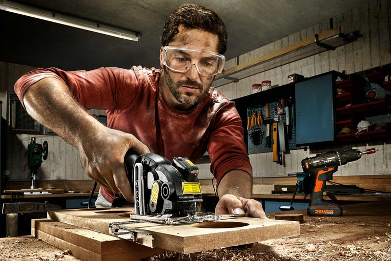 Mann arbeitet in einer Werkstatt mit einer Tauchsäge an einem Werkstück aus Holz. Eine Bohrmaschine ist im Hintergrund zu sehen.