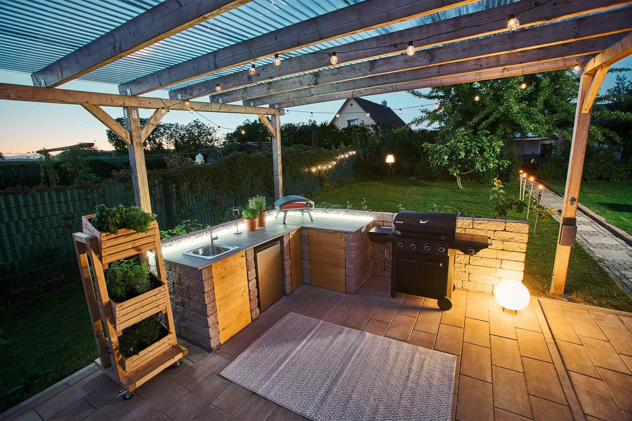 Moderne Außenküche unter einer Holzpergola mit Gasgrill, Schränken in Steinoptik, Holzfronten, Spüle und Pizzaofen auf gepflasterter Terrasse.