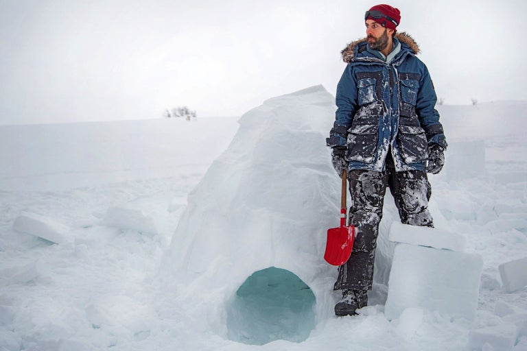 Mann in Winterkleidung steht neben einem Iglu mit einer Schaufel im Schnee.