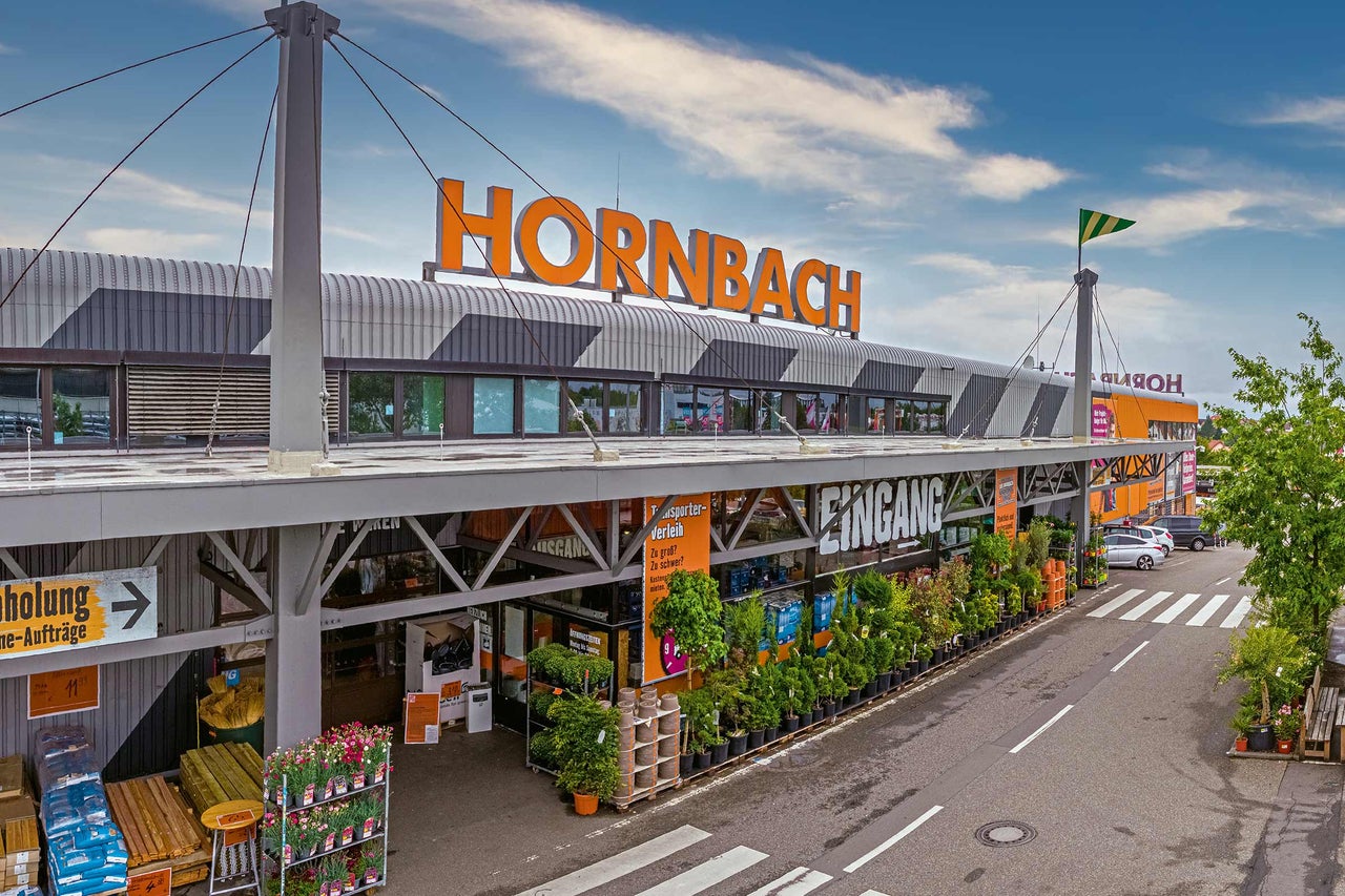  Außenansicht eines Hornbach Baumarktes mit Eingangsbereich und Pflanzen im Außenverkauf. 
