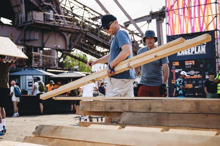 Zwei Personen tragen Holzbalken auf einem Festivalgelände bei Bauarbeiten für eine Holzkonstruktion.