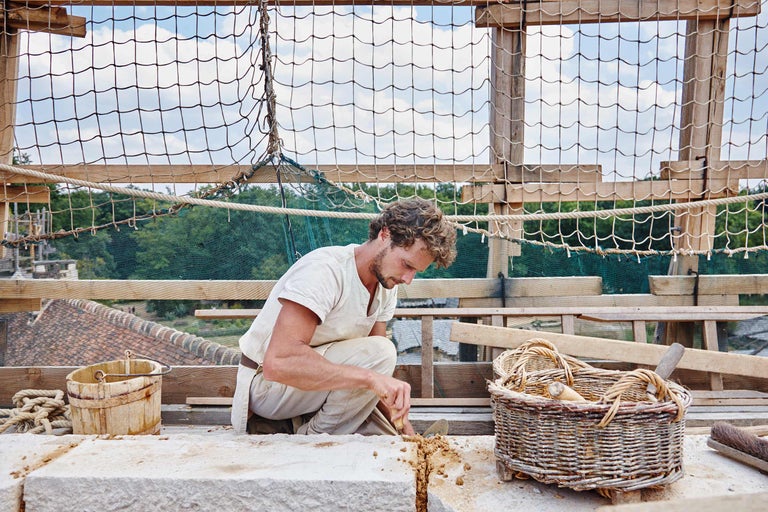 Ein Handwerker kniet auf einer Baustelle und bearbeitet Stein mit Werkzeugen. Im Hintergrund sind ein Holzeimer und ein Weidenkorb zu sehen.