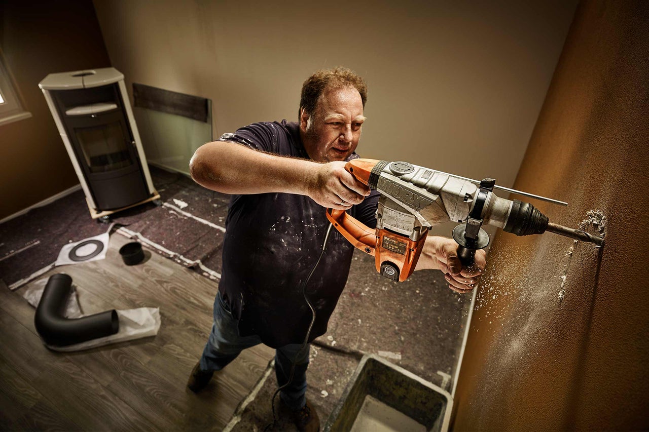 Ein Mann bohrt mit einem Bohrhammer ein Loch in eine Wand in einem Raum mit einem Ofen
