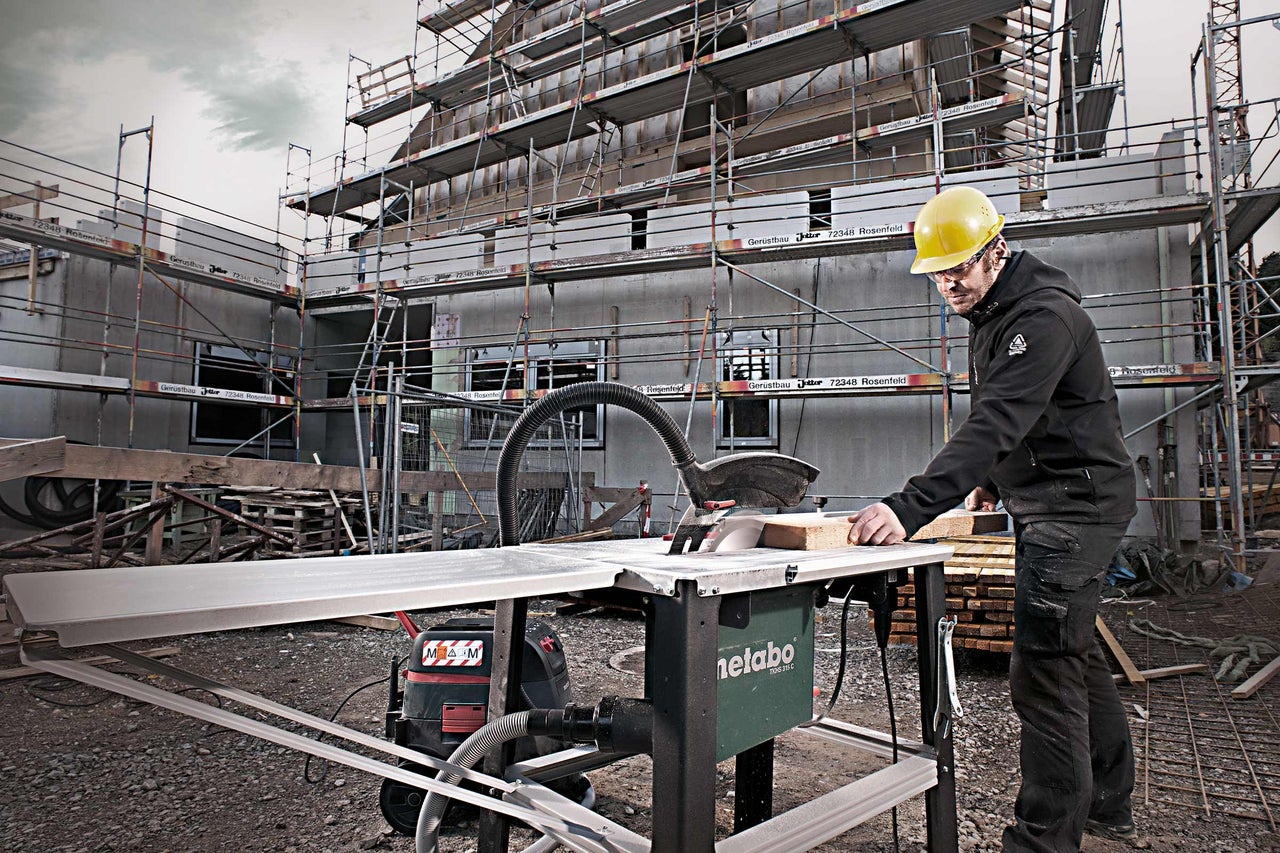 Ein Bauarbeiter mit Schutzhelm sägt mit einer Metabo Tischkreissäge ein Holzbrett auf einer Baustelle.