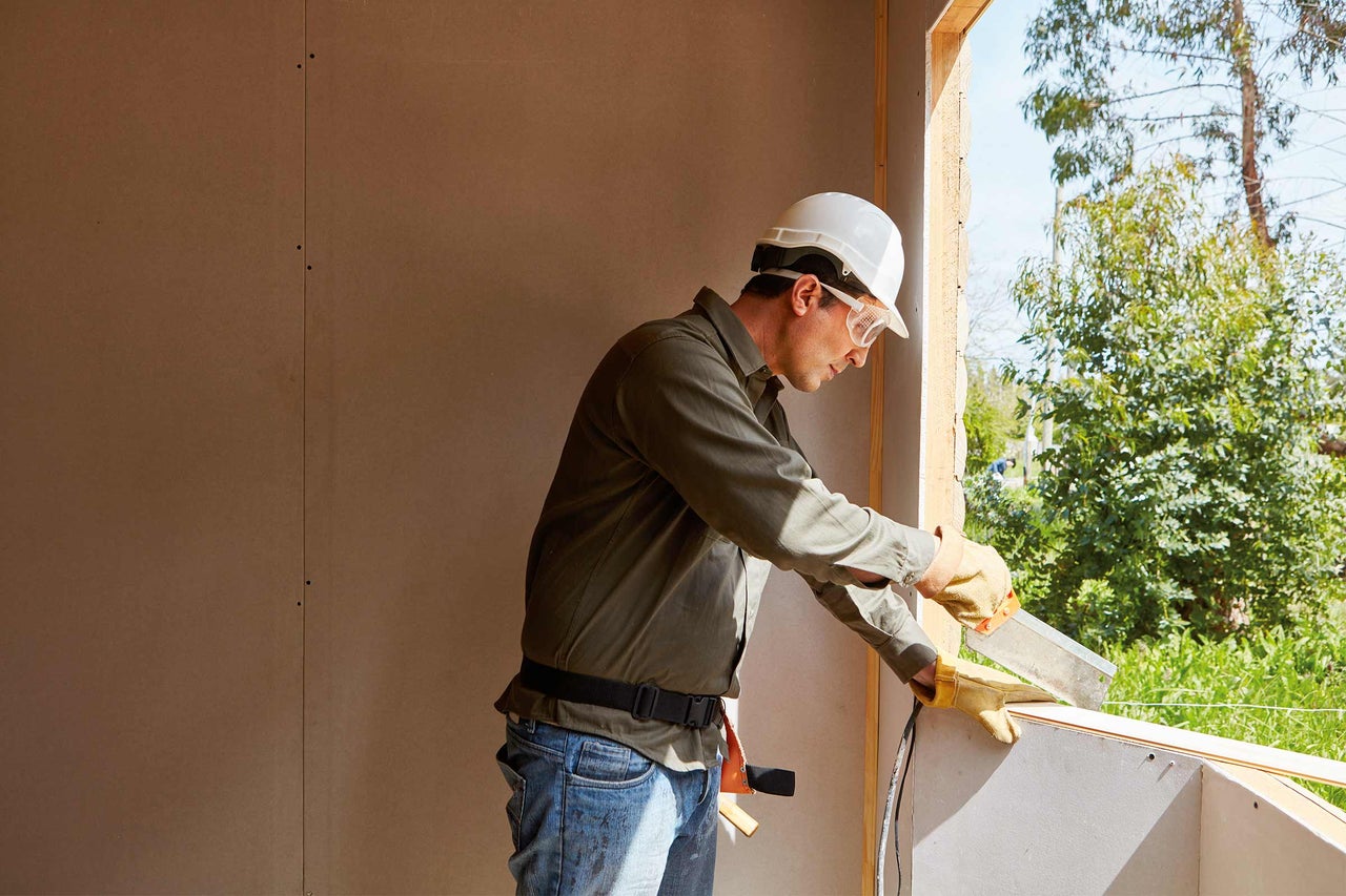 Ein Bauarbeiter mit Schutzhelm und Schutzbrille arbeitet an einem Fensterrahmen auf einer Baustelle.