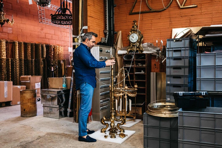 Ein Mann montiert einen goldenen Kerzenständer in einem Lagerraum mit Regalen, Kisten und anderen Gegenständen.