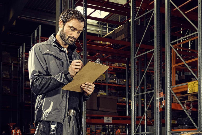 Ein Mann in Arbeitskleidung macht Notizen auf einem Klemmbrett vor Lagerregalen.