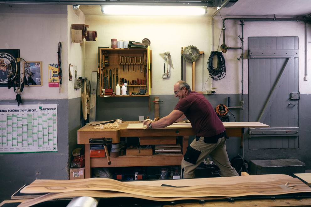 Klaus hobelt einen Holzkern an der Hobelbank. Vorn lieg Holzfurnier auf einem Tisch. Klaus hobelt einen Holzkern an der Hobelbank. Vorn lieg Holzfurnier auf einem Tisch.