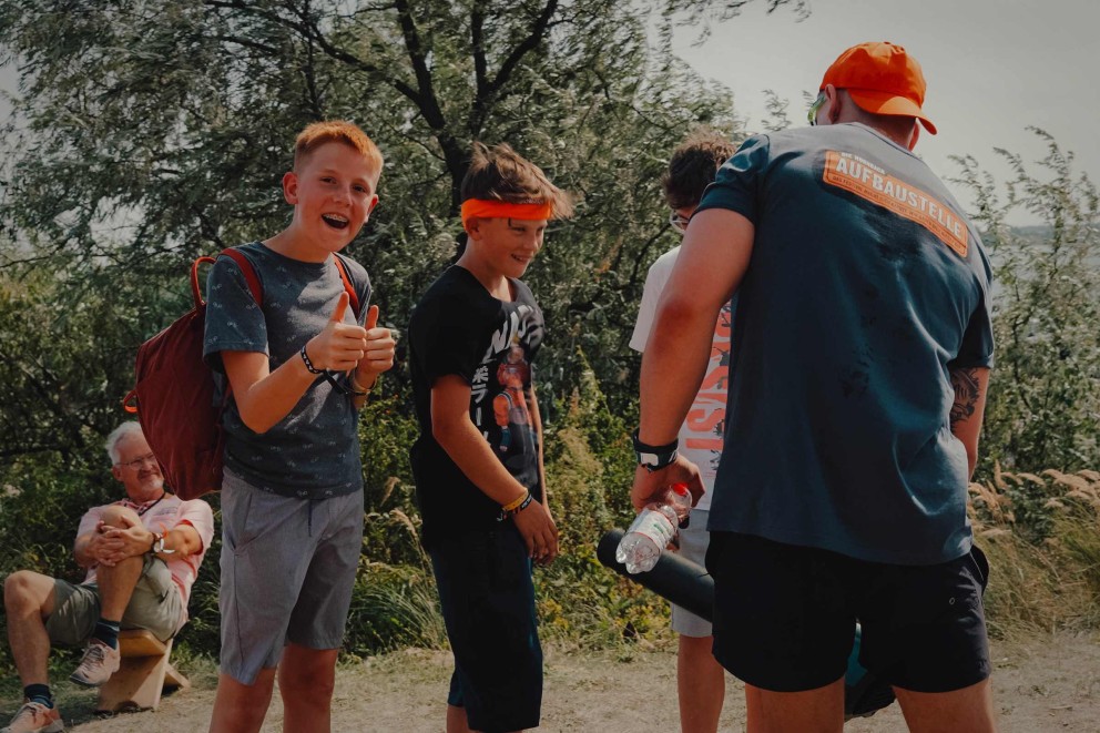Eine Gruppe von Menschen steht im Freien, darunter zwei Jungen mit Stirnbändern und ein Mann mit einer Wasserflasche, im Hintergrund ein Mann auf einer Bank und ein Baum.