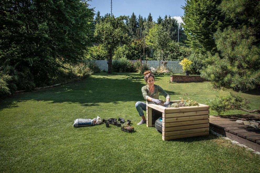 Eine Frau bepflanzt einen Hochbeetkasten im Garten, umgeben von Gartengeräten und Rasen.