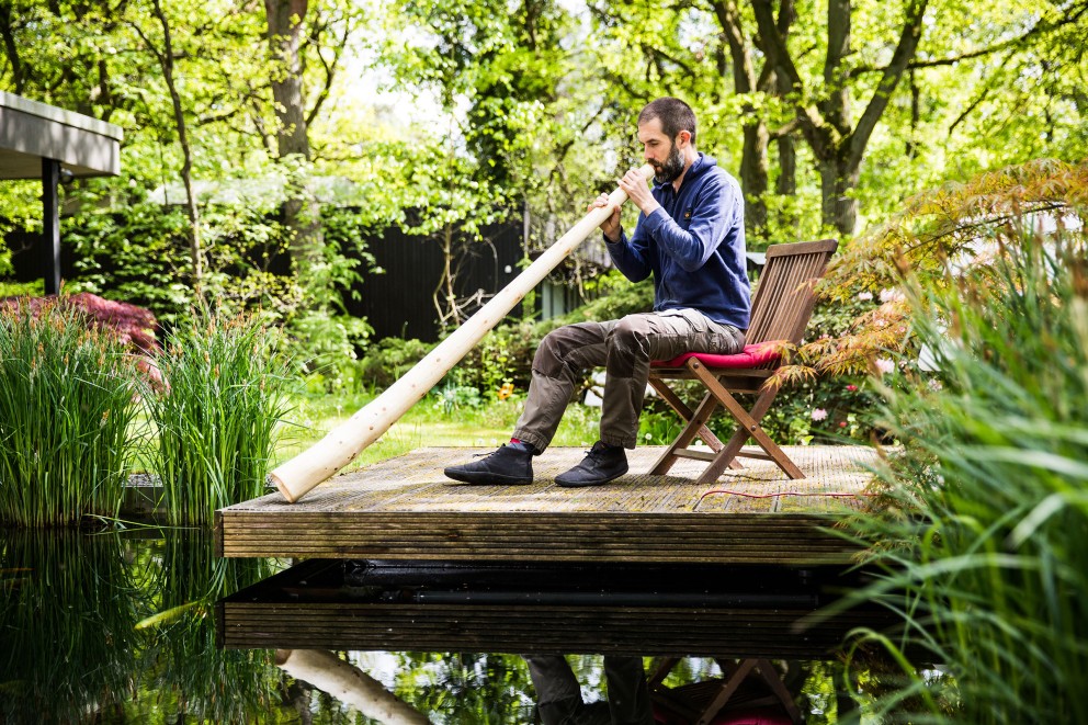Autor Sascha Borrée bläst Luft in sein selbst gebautes Didgeridoo, während er an einem Teich sitzt Autor Sascha Borrée bläst Luft in sein selbst gebautes Didgeridoo, während er an einem Teich sitzt