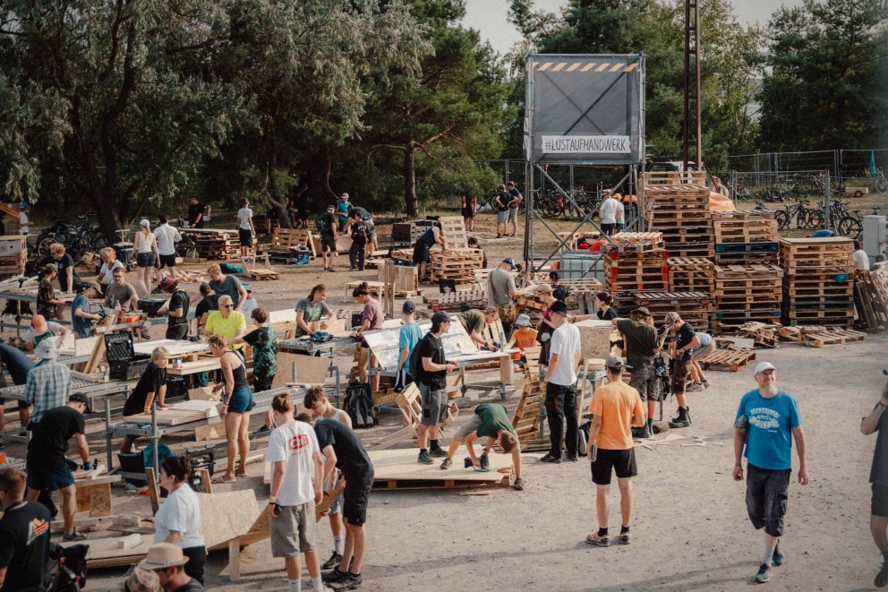 Eine Gruppe von Menschen arbeitet im Freien mit Holzpaletten und Werkzeugen, umgeben von Bäumen und einem Gerüst mit der Aufschrift '#LUSTAUFHANDWERK'.