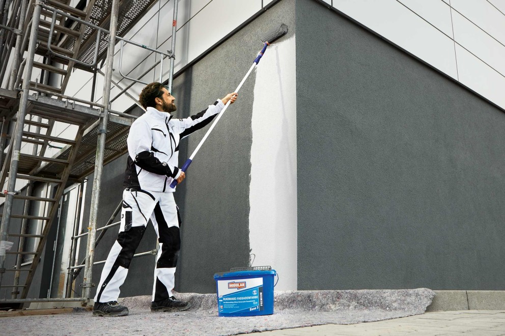 Ein Mann streicht eine Fassade mit einer Farbrolle und Fassadenfarbe. Im Vordergrund steht ein Farbbehälter der Marke Modulan.